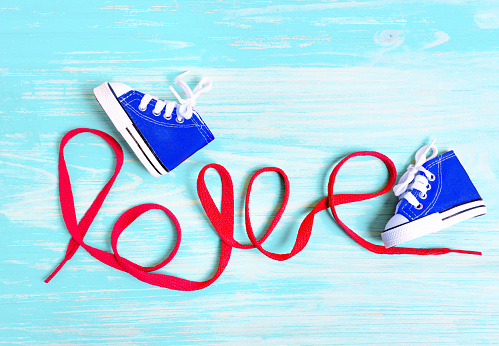 Miniature blue canvas shoes for toddlers and word LOVE made from a red shoelace on a blue wooden background. Kids fashion related concept.