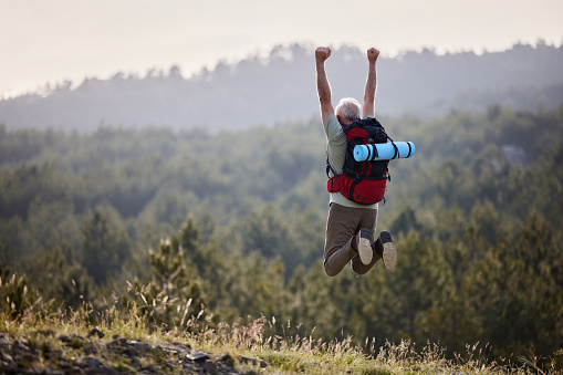 Rear view of a mature backpacker having fun while jumping with raised arms during hiking day in nature. Copy space.