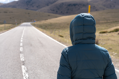 Unknown woman with hood looks ahead walking on totally lonely asphalt road with mountains background.