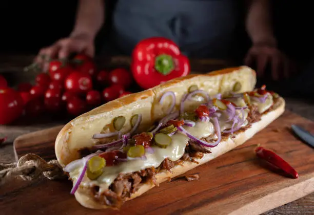 Delicious submarine sandwich with pulled turkey meat, melted cheese, pickles, red onions and barbecue sauce. Served on wooden board with decoration and chef with apron in the background.