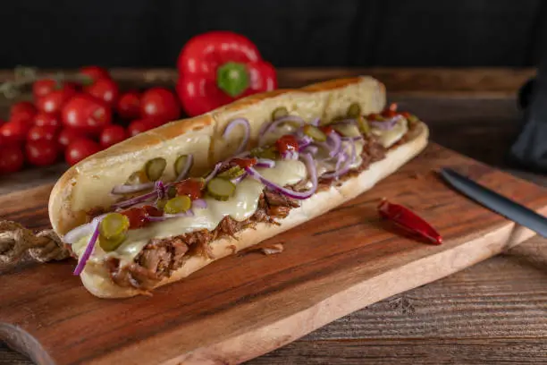Delicious homemade meat sandwich with pulled turkey, melted cheese, pickles, onions and barbecue sauce. Served ready to eat on wooden cutting board, closeup, front view