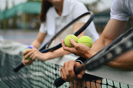 Cropped shot of male tennis coach hand holding balls, giving instructions to his student, standing by net at the court.