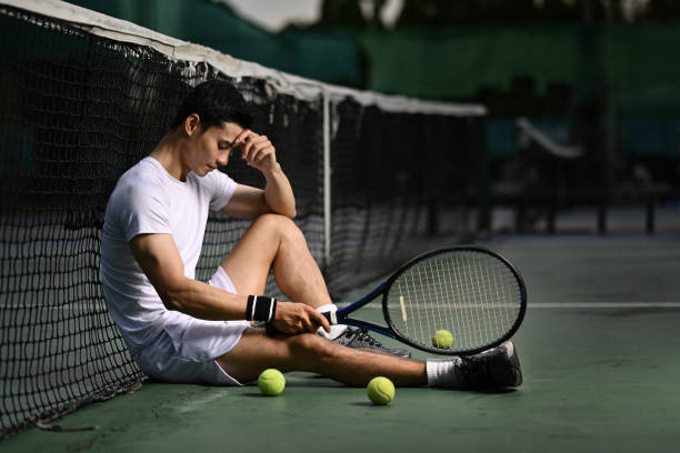 Side view of disappointed asian male tennis player sitting on the court, upset about competition loss, failure emotions. stock photo