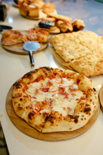 Neapolitan ham and cheese pizza with tomato base in the foreground with other pizzeria products blurred in the background of a counter