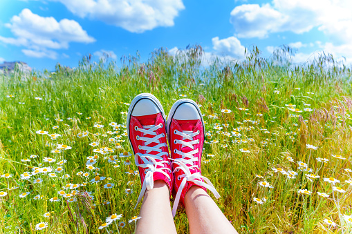 Pair of feet, adorned in red canvas shoes, lay amidst a vibrant field of daisies, with a picturesque view of the blue sky.
