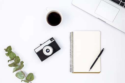 Minimalistic white background with retro camera, laptop, notebook, eucalyptus branch and cup of black coffee, top view, copy space.