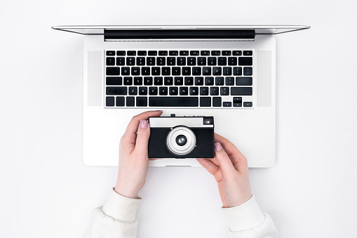 Retro camera in female hands and a laptop on a white background, top view. Minimalistic background, the concept of working as a photographer.