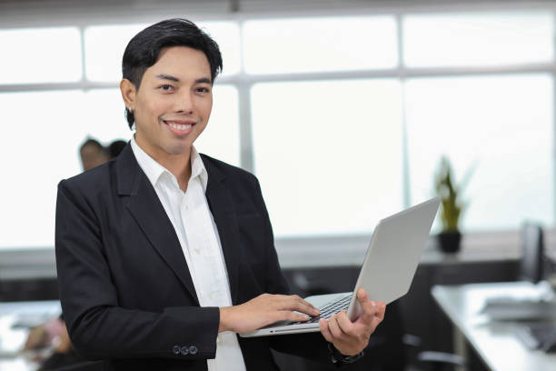 istock Attractive asian man employee worker in formal dress standing, using computer and looking at camera with coworker at workplace. Businessman smiling and working with notebook in the office. 1494753586