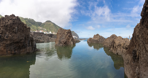 Madeira Island Portugal - 04 19 2023: Panoramic view of the natural pools on village of Porto Moniz, formed by volcanic rocks, village buildings in the background, coast of the Madeira island, Portugal