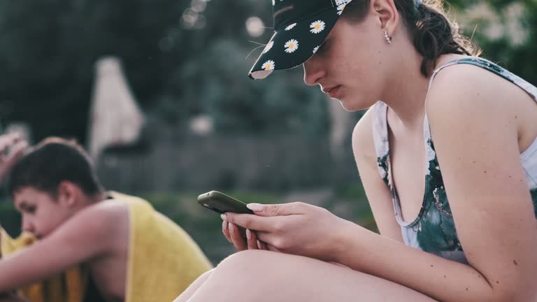 Beautiful Girl in a Bathing Suit Sits on a Sandy Beach with a Phone in Her Hands