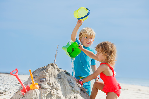 Close view of smiling boy and sister girl play with sand pour water from bucket on castle at the ocean beach during summer vacations