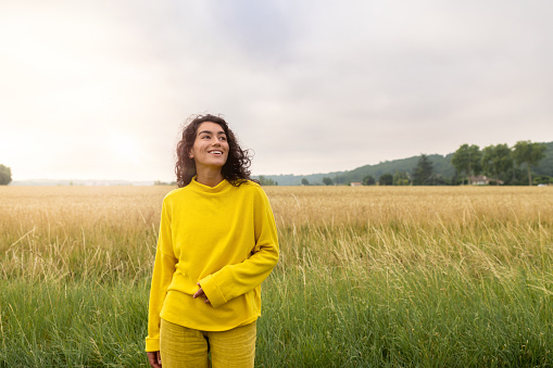 A medium wide angle view of a  female standing at golden hour in the early evening near Toulouse, France. She is dressed head to toe in yellow. She is smiling and looking away from the camera.