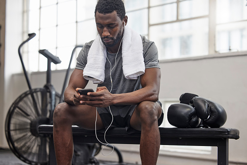 Phone, music and fitness with a black man taking a break in a gym during his boxing workout. Exercise, social media or sports app with a male boxer or athlete tracking progress and resting on a bench