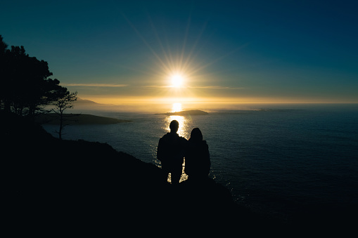 Silhouette of a young couple looking at the horizon on a cliff in Monteferro at sunset. Nigran - Spain