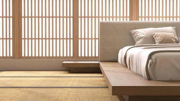 Wooden Japanese platform bed with bedside table, fabric headboard, brown blanket, pillow on tatami mat in sunlight from shoji window and leaf shadow