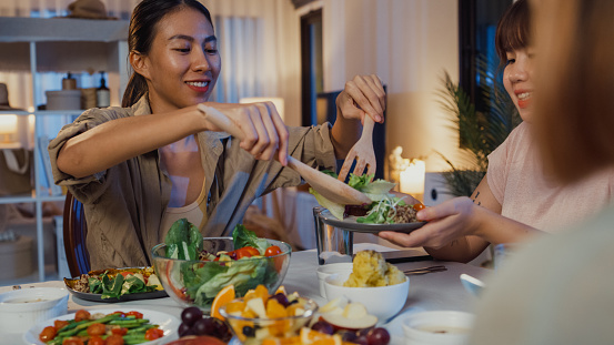 Young Asian women friends eating salad and talking fun sit at dining table in dining room at home at night. Lifestyle healthy food eating enjoying natural life and plant-based diet concept.