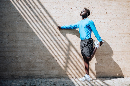 portrait of young black sportsman standing in front of wall and stretching