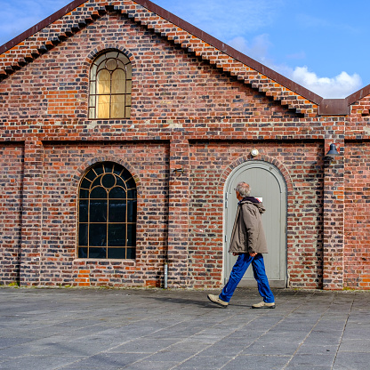 Sandnes, Rogaland, Norway, May 18 2023, Older Man Walking Past A Traditional Brick Building Under A Blue Sky In Evening Sunlight