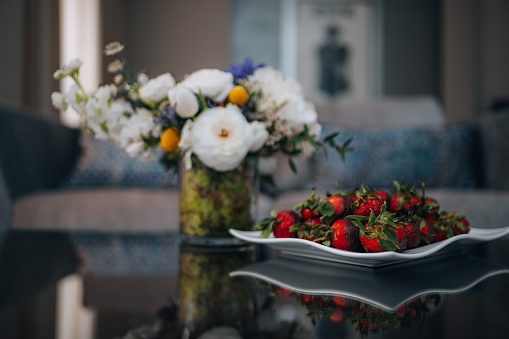 Fresh strawberries dipped in dark chocolate next to a vase with a white flower bouquet in a cozy room