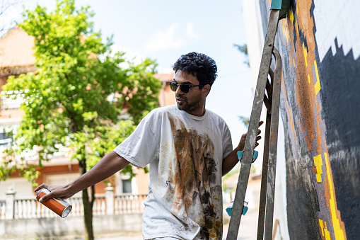 Street artist working on colored graffiti at public space wall - Modern art perform concept of urban guy painting live murales with yellow and orange aerosol color spray - Bright sunflare filter