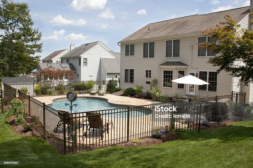 Lush backyard pool and patio behind colonial style home. Lush, resort-like backyard salt water swimming pool with flagstone stamped concrete patio behind colonial style home.  Bronze fencing is seen around the pool area of this suburban backyard. Fence Stock Photo