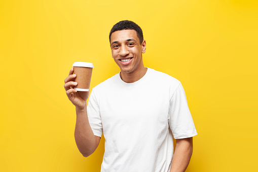 young african american guy in white t-shirt holds and recommends coffee in paper cup on yellow isolated background, man points and advertises cup of drink