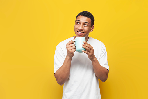 young african american guy in white t-shirt holds cup of coffee on yellow isolated background, man with drink looks at copy space