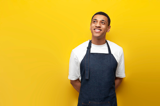 young African American barista guy in uniform on yellow background looks at copy space, delivery service worker
