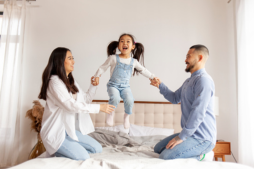 happy asian family with daughter play and rejoice at home on the bed, little active korean girl jumps on the bed and screams