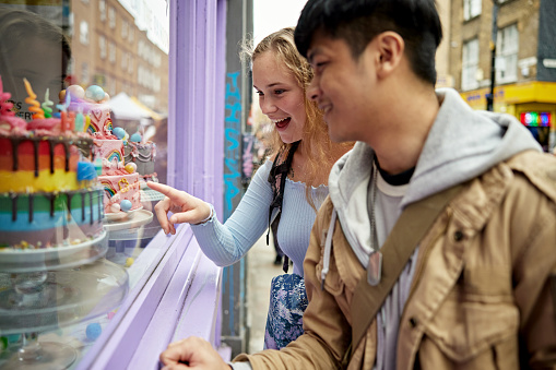 Casually dressed Londoners in early 20s walking then pausing to point and smile at multicolored decorated desserts.