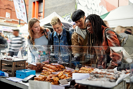 Waist-up view of Asian, Black, and Caucasian friends in late teens and early 20s talking and smiling as they admire options for lunch.