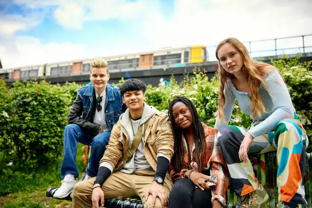 Front view of young Caucasian, Asian, and Black friends, casually dressed, sitting together on bench and smiling at camera with train passing in background.