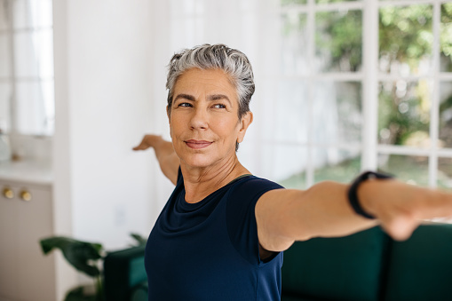 Active senior woman doing the warrior pose at home, she stretches her arms with a confident look on her face. Mature woman practicing yoga to improve her strength, balance and stability.