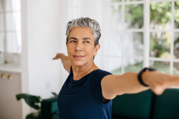 Senior woman stretching her arms in powerful warrior yoga pose Active senior woman doing the warrior pose at home, she stretches her arms with a confident look on her face. Mature woman practicing yoga to improve her strength, balance and stability. warrior position stock pictures, royalty-free photos & images