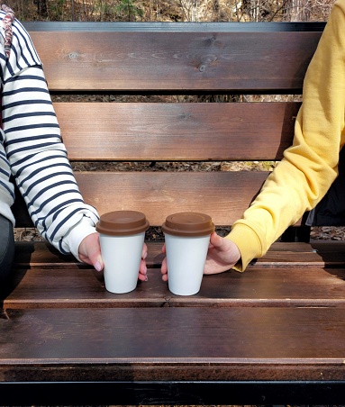 A woman's and men's hand hold coffee mugs on a park bench.