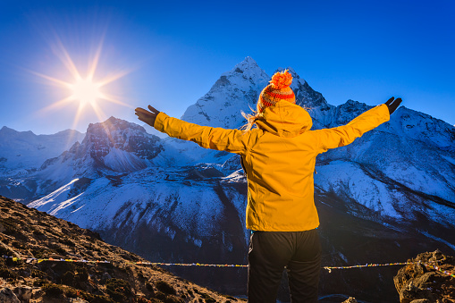 Young woman standing on the top of a mountain and watching sunrise over Himalayas, she is wearing yellow jacket, Her arms are outstretched. The sun and mount Ama Dablam on a background. Mount Everest National Park. This is the highest national park in the world, with the entire park located above 3,000 m ( 9,700 ft). This park includes three peaks higher than 8,000 m, including Mt Everest.