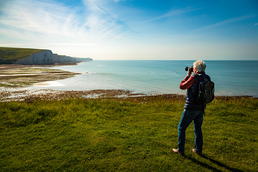 A senior man using his professional camera to shoot images of the landscape on top of the cliffs overlooking the sea at Seven Sisters cliffs in southern England.