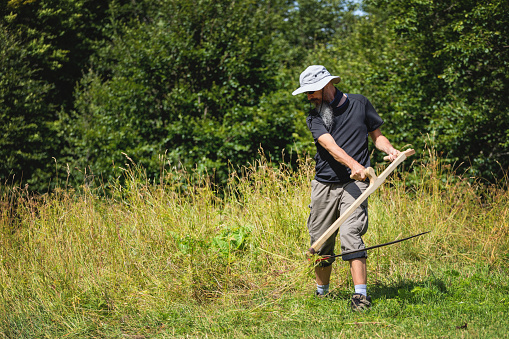 Mid adult man mowing grass at high mountan plain at summer using scythe