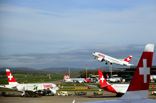 Zurich / Kloten, Switzerland: Swiss Bombardier CSeries CS100 Airbus A220 (HB-JBA, MSN 50010), taking off at Zurich Airport (ZRH), other aircraft on the ground (Swiss, Helvetia, Air Serbia) - Swiss International Air Lines is the flag carrier of Switzerland, with headquarters and hub at Zurich Airport (legal headquarters  in Basel), it is part of the Lufthansa Group and a member of Star Alliance