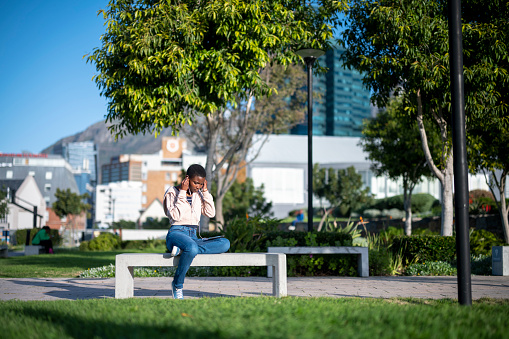 Young African woman sitting on a bench in an open park putting on her earphones to connected to her mobile phone