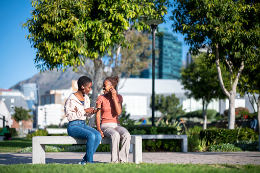 Two happy African girls sitting on a park bench sharing earphones to listen to music on a mobile phone