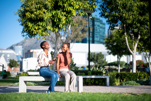 Two happy African girls sitting on a park bench on a clear day sharing earphones laughing as they listen to something amusing on a mobile phone