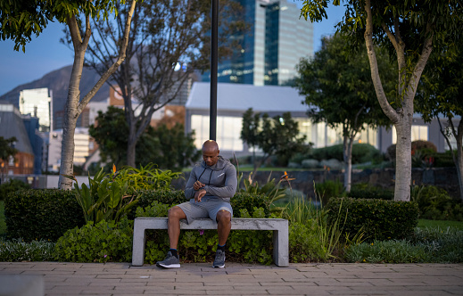 Man wearing activewear rests on a park bench  after his jog & looks at his smartwatch