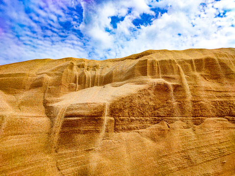 Sand crumbles down the slopes of a sandy mountain under a blue sky with clouds - photo of a sand dune against a blue sky with white clouds. Small grains of sand. Dune background. The wind blew away the sand. Clouds in the sky. Mountain of sand and the sky. Sand waterfall.