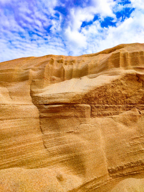Layers of sand in a sand mountain and a blue sky with white clouds - photo of a sand dune against a blue sky with white clouds. Small grains of sand. Dune background. The wind blew away the sand. Clouds in the sky. Mountain of sand and the sky. Vertical photo.