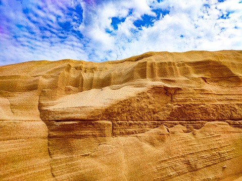 A sand mountain with layers of sand against a blue sky with clouds - photo of a sand dune against a blue sky with white clouds. Small grains of sand. Dune background. The wind blew away the sand. Clouds in the sky. Mountain of sand and the sky.