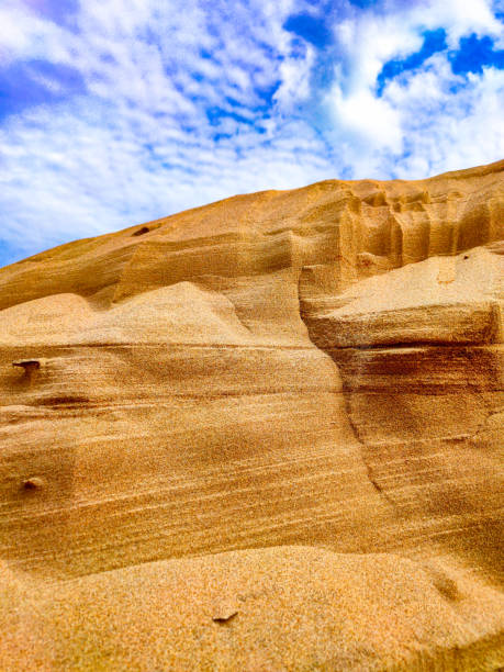 A mountain of layers of sand under a blue sky with clouds - photo of a sand dune against a blue sky with white clouds. Small grains of sand. Dune background. The wind blew away the sand. Clouds in the sky. Mountain of sand and the sky.