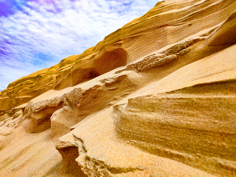 Sandy mountain against a blue sky with clouds - photo of a sand dune against a blue sky with white clouds. Small grains of sand. Dune background. The wind blew away the sand. Clouds in the sky. Mountain of sand and the sky.