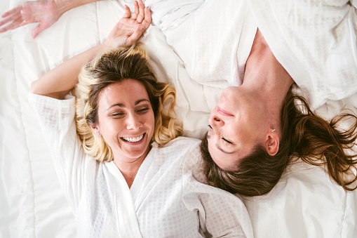 Happy two friends Caucasian young women in white bathrobes enjoying and looking at each other together with smiles lying on the bed. LGBTQ couples fun leisure activities spa beauty natural treatment