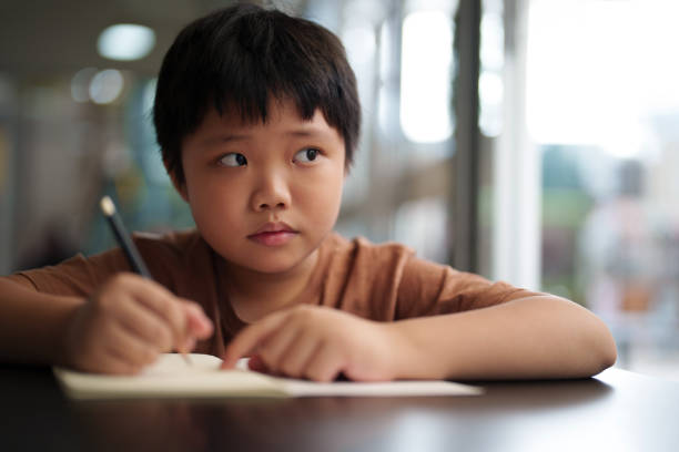 A close-up shot captures an Asian boy deep in thought, looking away, while using a pencil to write and draw on a white book
